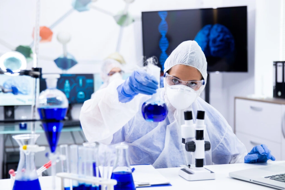Female scientist in protection equipment holding and looking at a test tube with blue solution