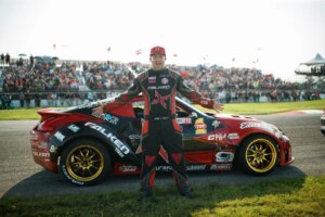 professional drift driver Jeff Jones standing next to his red drift car on a racetrack sponsored by top 1 oil