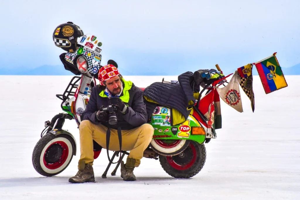 A man wearing a bandana, gloves, and a jacket sits next to a heavily customized, vespa adorned with flags and stickers